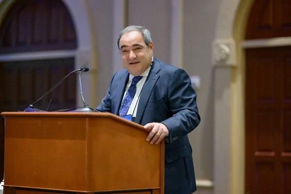 Edgar Romano standing behind a podium, wearing a suit and tie, speaking at the WILG annual convention in California. Romano received the Lifetime Achievement Award.