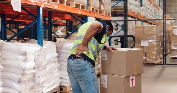 A warehouse worker in a high-visibility vest experiencing back pain, clutching his lower back while leaning on stacked boxes.